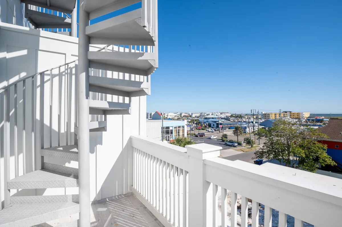 A striking spiral staircase leads up to an open balcony with a view of the waterfront and neighboring buildings. The scene feels optimistic and social, inviting guests to imagine relaxing at sunset or enjoying social time outdoors with panoramic views.