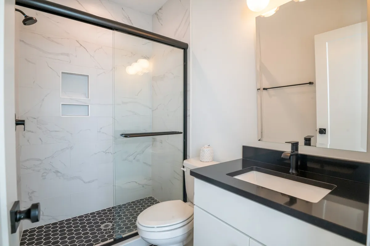 A bright, modern bathroom with a glass shower enclosure, black countertop, and stylish hexagonal floor tiles. The atmosphere is serene and sophisticated, ideal for relaxing and self-care routines.