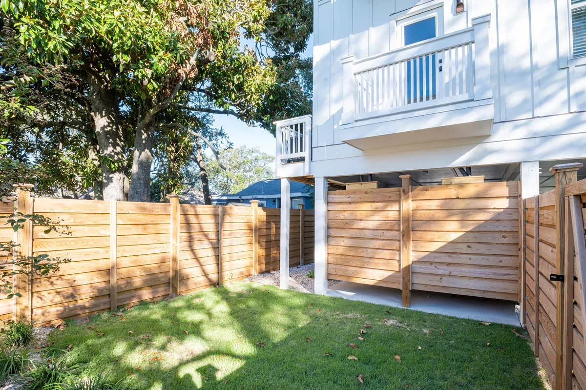 A peaceful, sunlit backyard with a wooden fence, green grass, the exterior of the house, and a shady tree canopy. The space looks inviting for both children and pets to play, or for adults to relax outdoors. Visible is ample space for outdoor activities, meals, and relaxation.