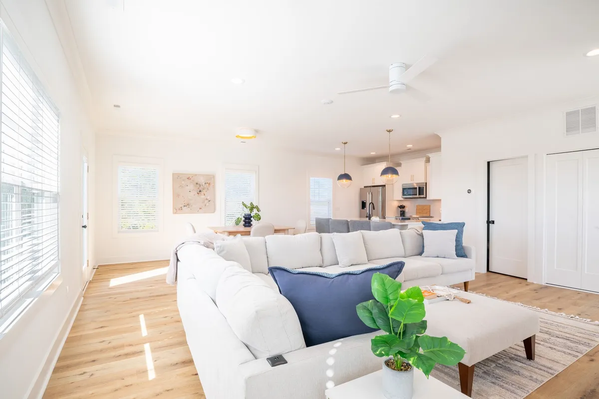 The image showcases a spacious, open-concept living area with a large, comfortable sectional sofa, a modern dining area, and a kitchen island—all bathed in natural light from expansive windows. The room exudes a welcoming, family-friendly atmosphere, perfect for gatherings and relaxation.