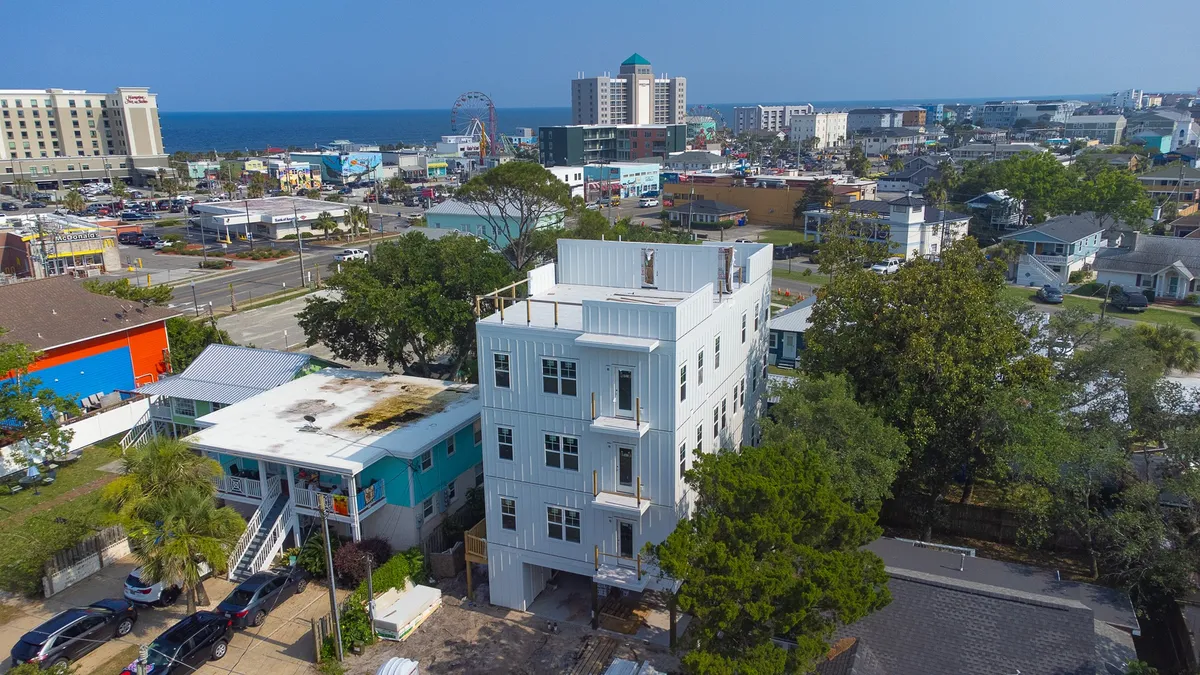 A vibrant exterior shot showcasing the large white building with the ocean in the background, the amusement park, and colorful buildings nearby. The scene is alive with energy, highlighting the proximity to the boardwalk, amusements, and the beach itself.