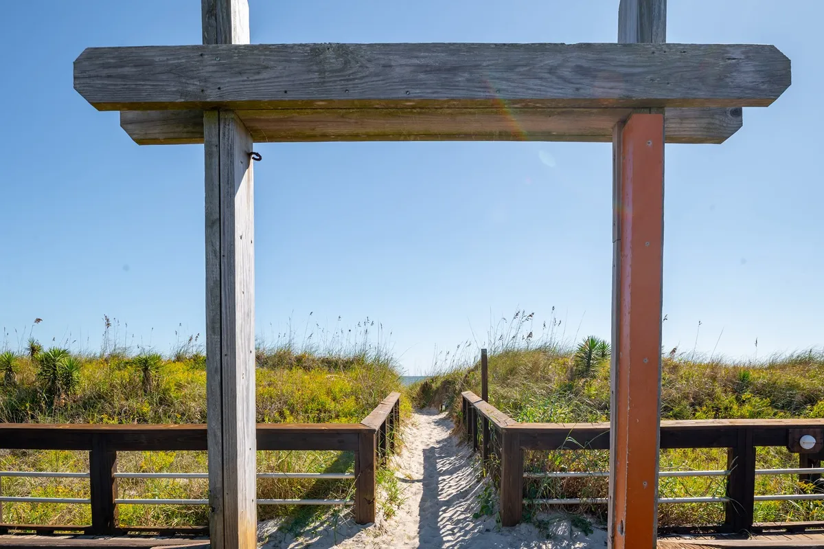 A serene pathway winds through a wooden archway bordered by green vegetation, leading directly to a sandy beach. The atmosphere is calm and inviting, evoking a sense of nature and adventure.