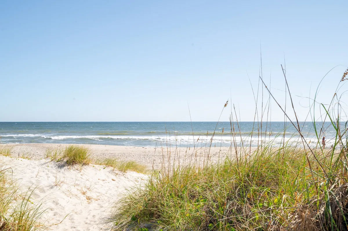 A serene beachfront view with sand dunes, ocean waves, sea grass, and a bright blue sky. The scene evokes classic beach activities like sunbathing, walking, and sandcastle building, perfect for family fun just steps away.