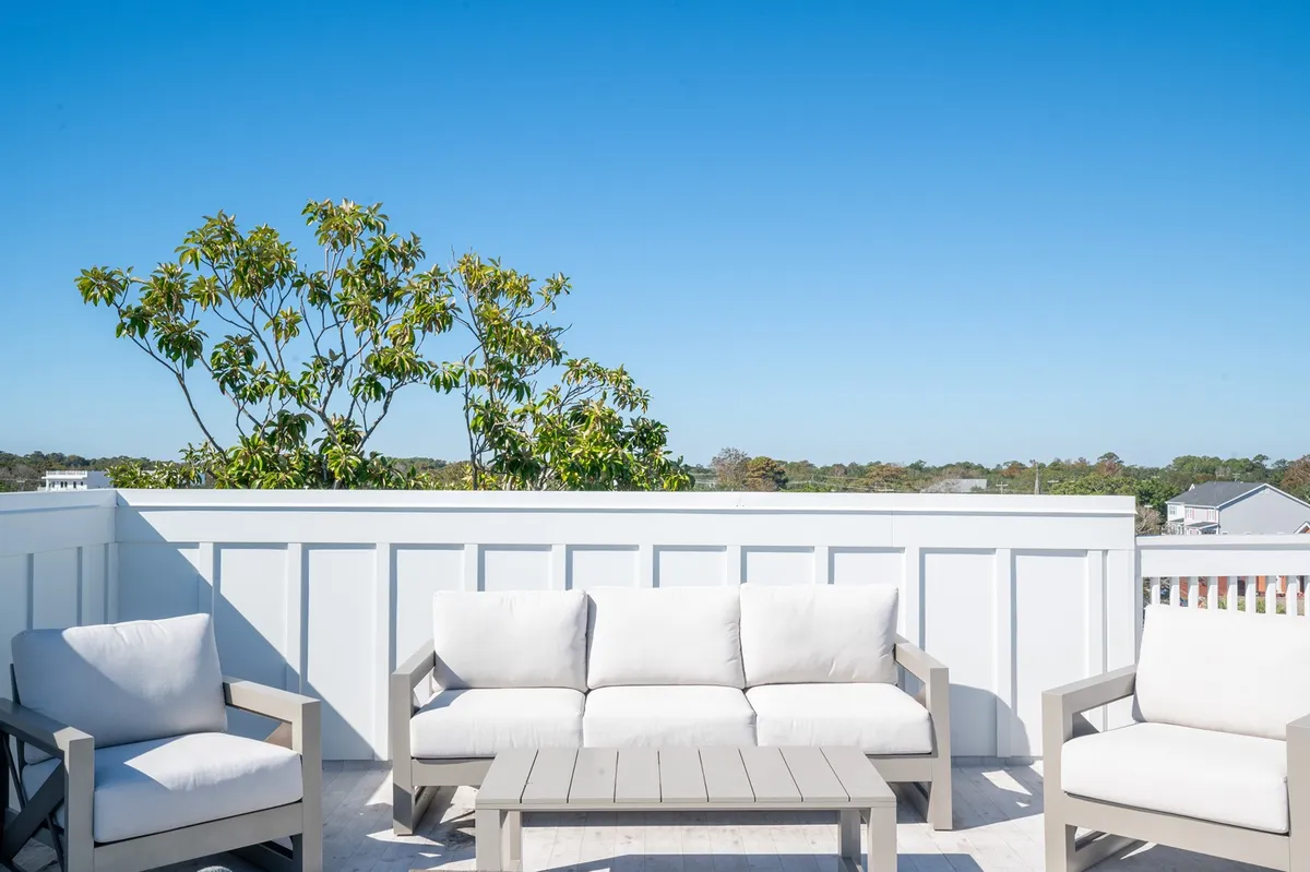 An inviting outdoor seating area is framed by lush green trees and a clear blue sky, suggesting a tranquil spot for sunset cocktails or morning coffee. The space feels private yet open, echoing the rooftop deck experience described in the content.