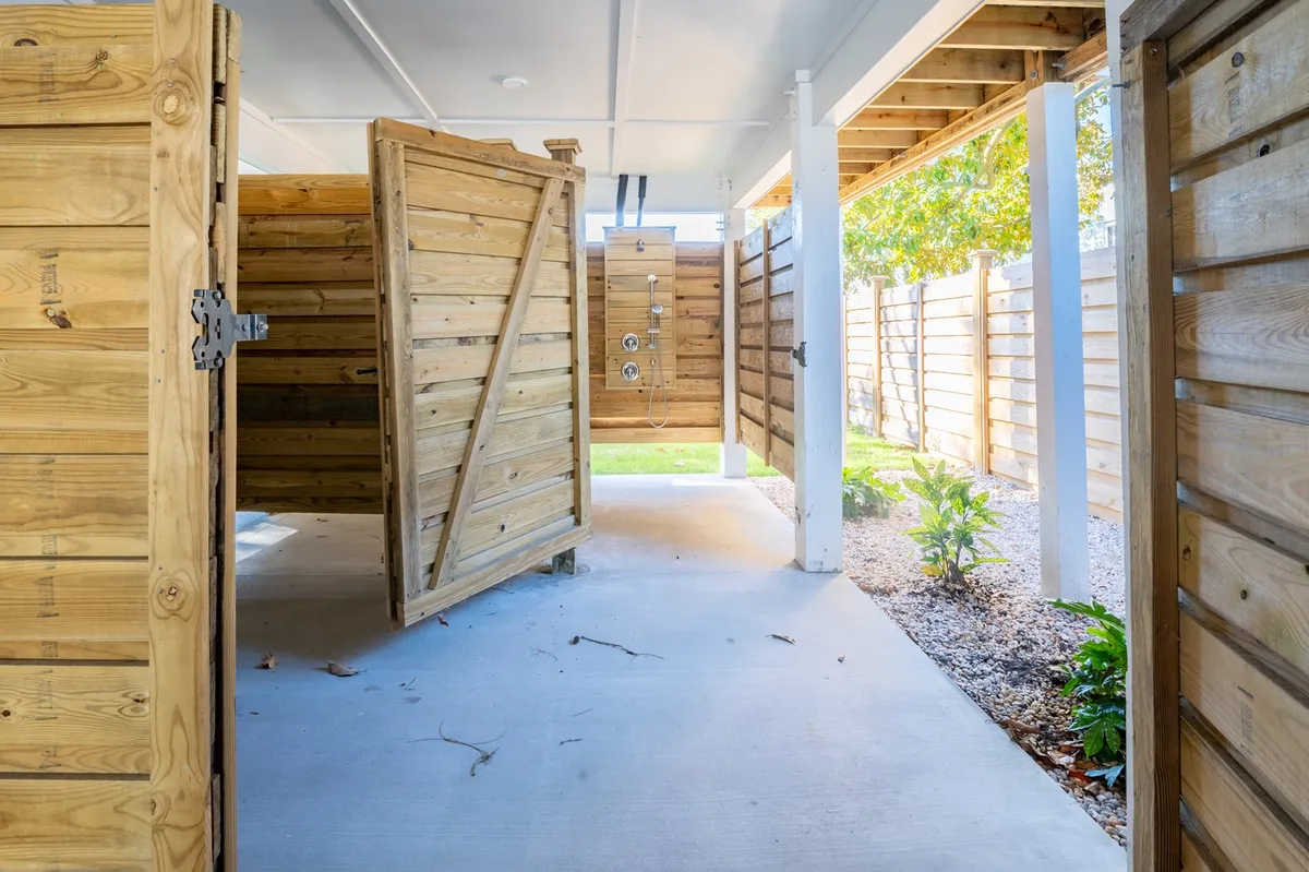 Outdoor wooden shower stalls set along a concrete path and surrounded by lush greenery, suggesting a refreshing, private spot for rinsing off after the beach. The natural setting adds to the relaxed, easygoing mood.
