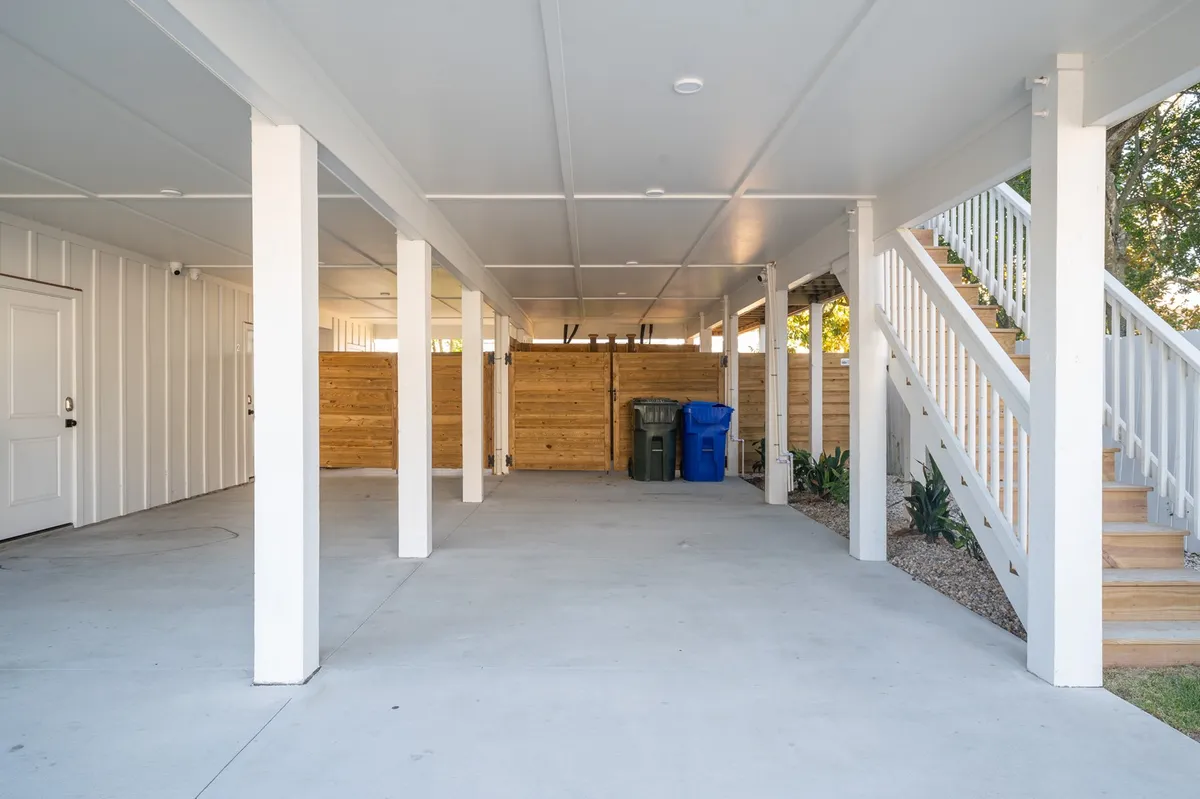 A shaded ground-level area beneath the house with supporting columns and visible stairs leading upwards. There’s a wooden fence and space for parking vehicles or storing beach gear, and the area invites casual gatherings or relaxation after returning from the beach.