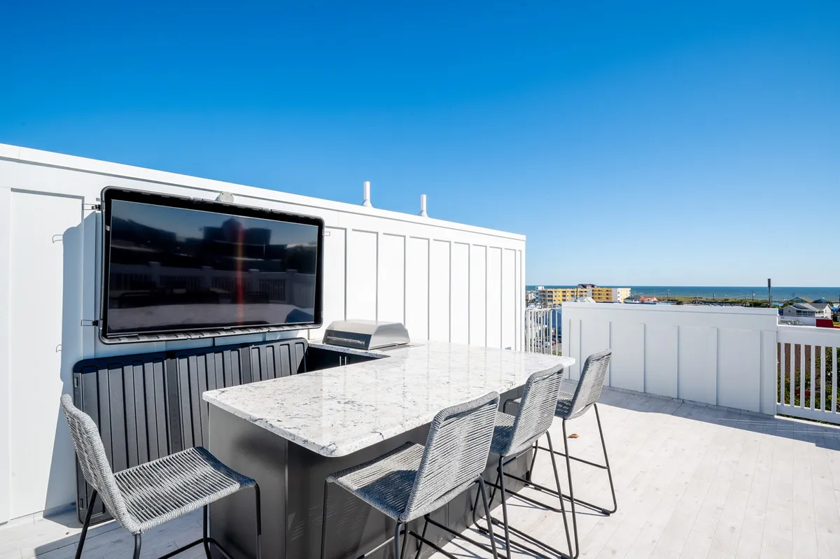 A stylish rooftop deck featuring an outdoor bar, flat-screen TV, and panoramic ocean views. Plush seating invites guests to relax, while a grill station hints at lively gatherings. The sun sets over the water, casting a golden glow across the social space.