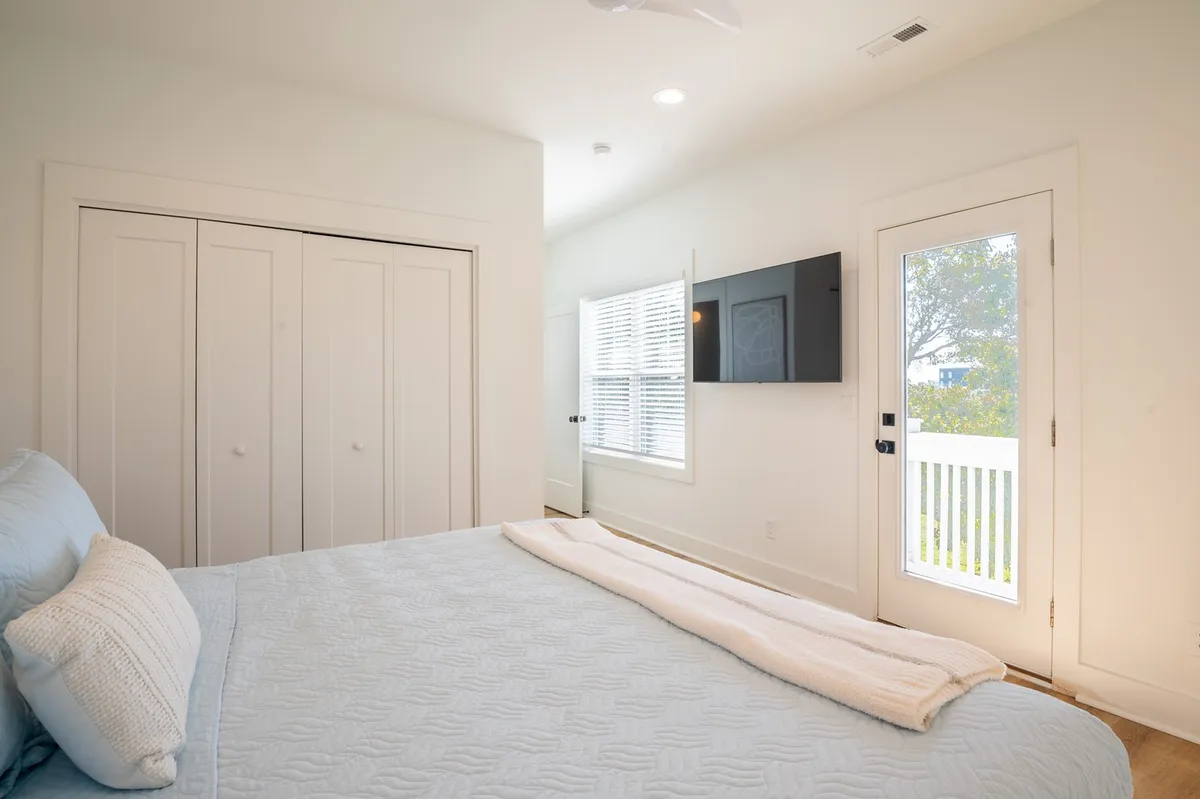 A serene bedroom with a king-sized bed featuring light blue bedding, a window with white shutters letting in natural light, a wall-mounted TV, and a door leading out to a private balcony. The space feels tranquil and welcoming, perfect for restful nights and leisurely mornings.